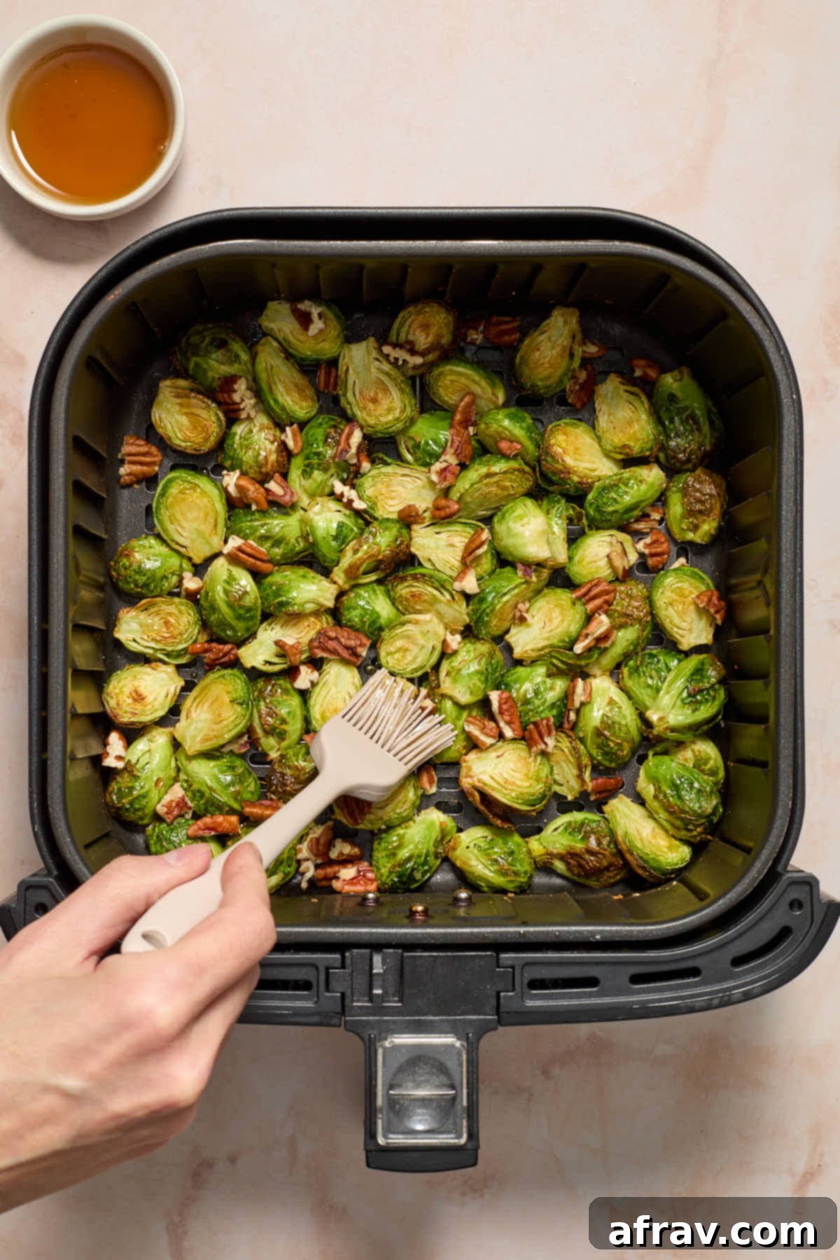 Pecans added to the Brussels sprouts in the air fryer basket, with maple syrup being brushed on, showing the final flavoring step.