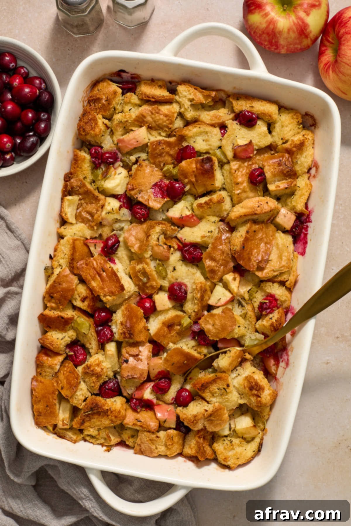 Festive Apple Cranberry Stuffing 2 Stuffing in a white baking dish with a spoon inserted into it, and cranberries and apples on the side.
