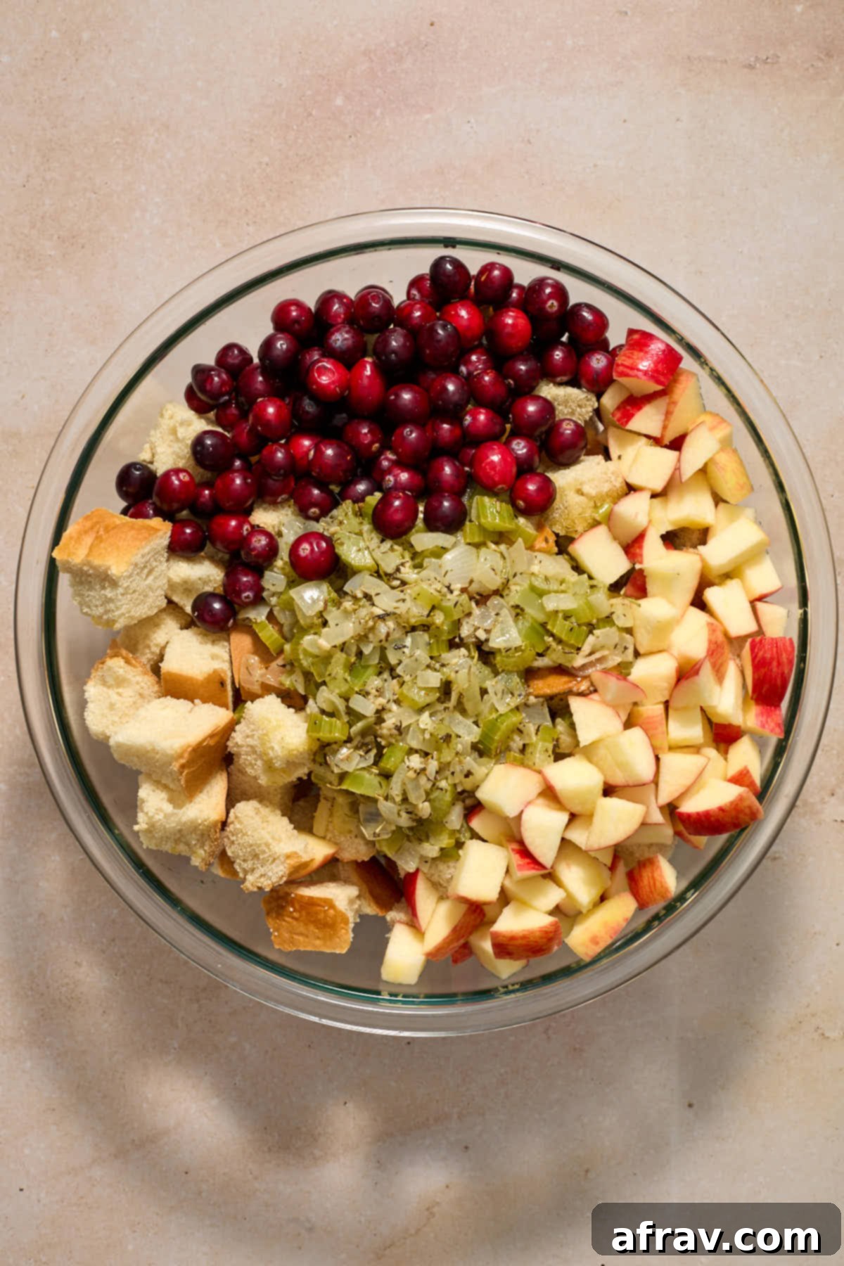 Festive Apple Cranberry Stuffing 8 Bread cubes, vegetable mixture, chopped apples and cranberries added to a large glass bowl.