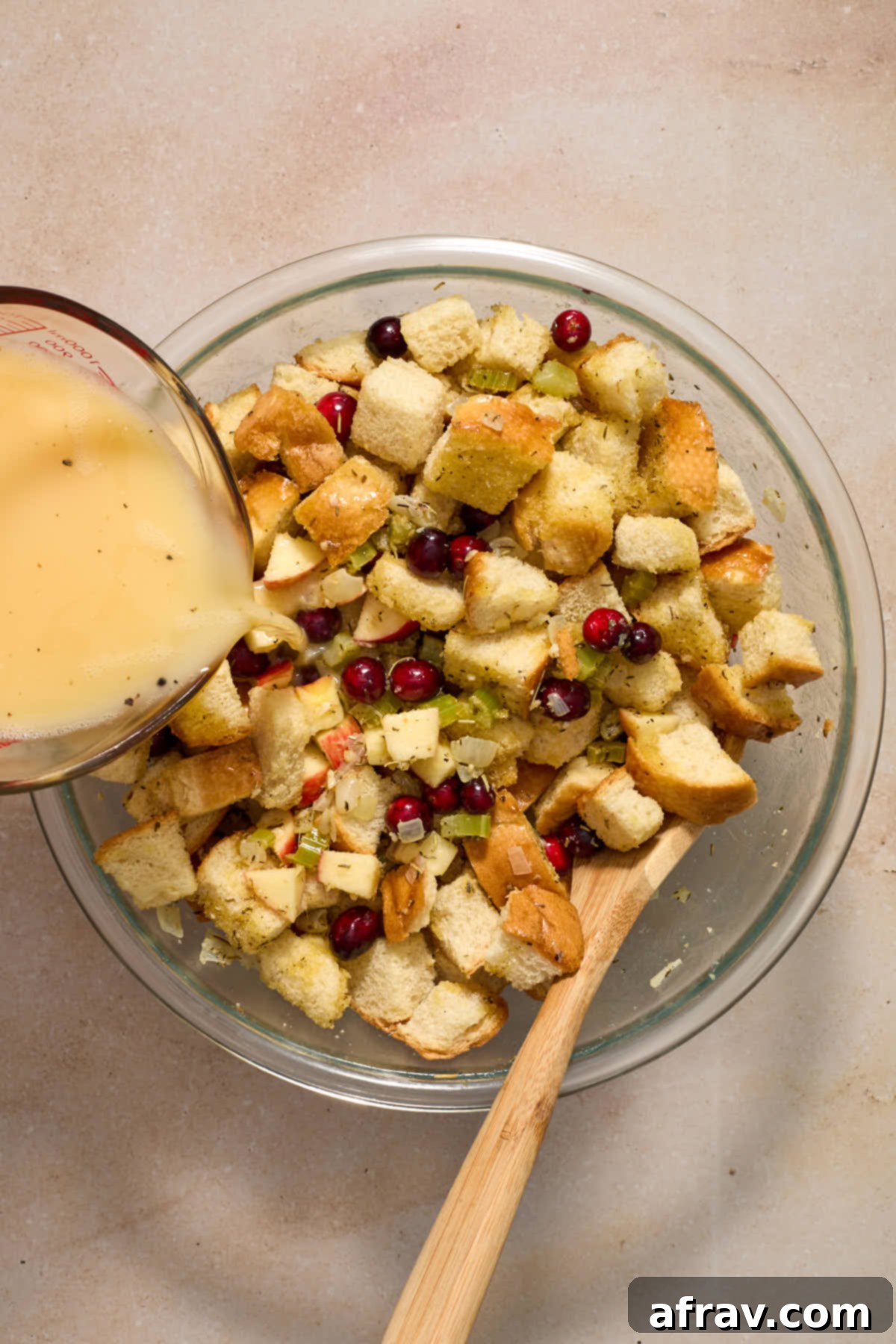 Festive Apple Cranberry Stuffing 9 Stuffing ingredients mixed together in a glass bowl with a wooden spoon and egg/broth mixture being poured over it.