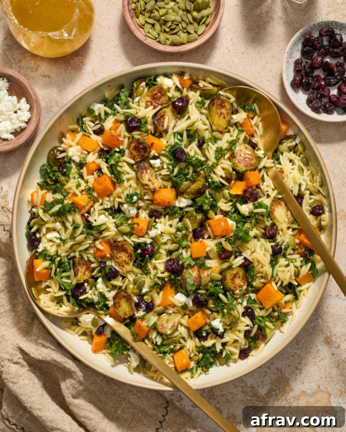 A vibrant Fall Orzo Salad displayed in a large, shallow bowl, accompanied by serving spoons and an arrangement of the salad's colorful toppings.
