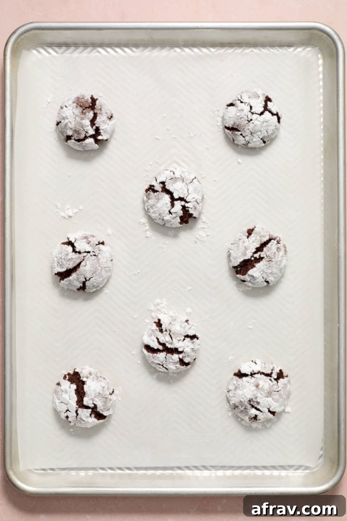 Freshly baked cookies on a parchment-lined baking sheet, showcasing their perfectly formed cracks and dusted with powdered sugar.